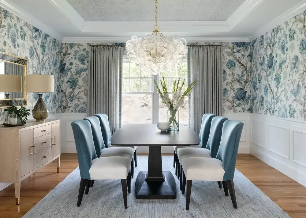 Dining room with blue and cream floral wallpaper above white wainscoting and a glass chandelier